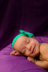 Sleepy smiling baby girl with turquoise ribbon on her head; purple background