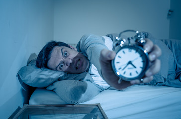 Young man in bed with alarm clock feeling desperate and distress not able to sleep with insomnia
