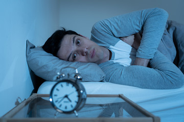 Portrait of young man in bed staring at alarm clock trying to sleep feeling stressed and sleepless