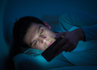 Young caucasian sleepless man bored in bed surfing on the Internet addicted to mobile phone