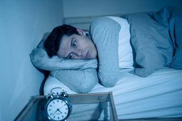 Portrait of young man in bed staring at alarm clock trying to sleep feeling stressed and sleepless
