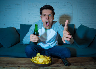 Portrait of excited young man on couch watching football at night drinking beer. Sports fans concept