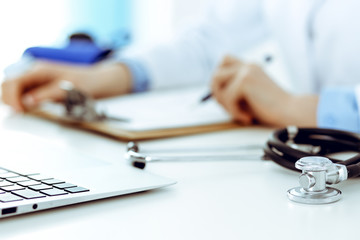 Closeup of stethoscope. Female doctor fills up medical form while sitting at the desk in hospital. Healthcare, workplace and cardiology in medicine concept