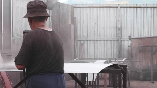 Old man worker applying anti-corrosion powder coating to metal sheet in slow motion. He is using spray gun in factory. Stands back to camera and does his job. Protection metal sheet from destruction.