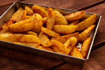 Potato wedges, oven baked, in a baking tray on a dark rustic wooden background