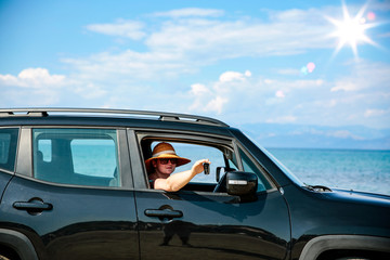 A woman wearing sunglasses and a hat sitting  in a summer black car on sandy beach view.