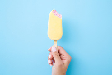 Young woman hand holding pink ice cream with white chocolate glaze on pastel blue background. Bitten food. Closeup.