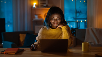 Portrait of Beautiful Black Girl Uses Computer while Sitting at Her Desk at Home, She's Wearing Warm Sweater. In the Evening Creative Woman Works on a Computer In Her Cozy Living Room.  - obrazy, fototapety, plakaty