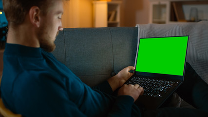 Fototapeta na wymiar Man Sitting Relaxes on a Couch Works on a Laptop with Green Chroma Key Screen. Late at Night in His Living Room Man Uses Notebook Computer. 