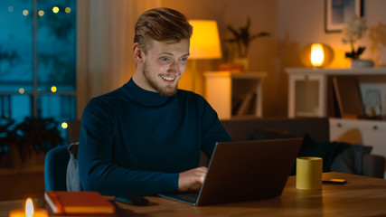 Handsome Smiling Man Works on a Laptop while Sitting at His Desk at Home. Portrait of a Young Freelancer Works on Computer in His Cozy Living Room with Warm Evening Lighting in Background.