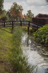 Fototapeta premium Pretty rural buildings with wooden bridge and trees. Idyllic countryside landscape. Typical holland houses with vintage bridge over river. Old buildings in Zaanse Schans, Netherlands. 