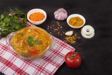Red lentil soup with tomatoes, herbs, onions, garlic and spices in a glass plate standing on a tablecloth on a black background.