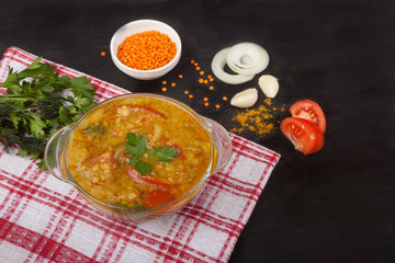 Red lentil soup with tomatoes, herbs, onions, garlic and spices in a glass plate standing on a tablecloth on a black background.