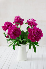 Purple garden peonies in a white enamel jug on a white wooden background, rustic style