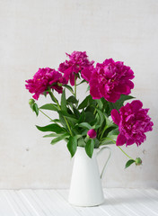 Purple garden peonies in a white enamel jug on a white wooden background, rustic style