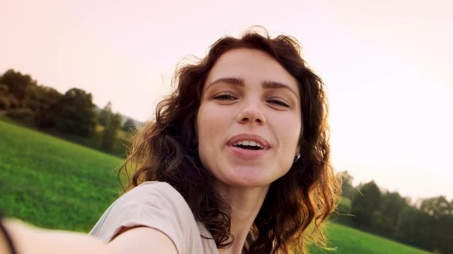Young woman with curly hair makes selfie and send a kiss at sunset on the background of a beautiful summer landscape.  Woman shoots herself