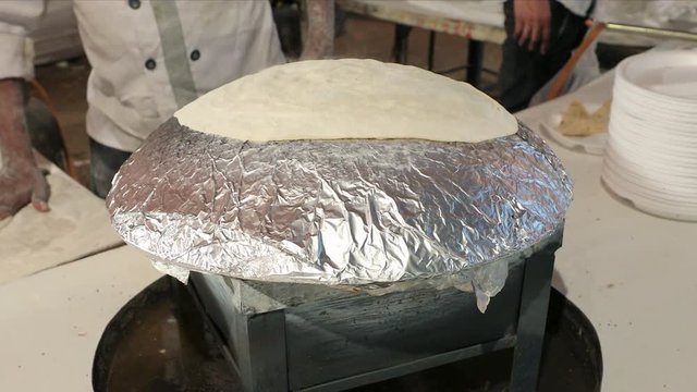 A uniformed man cooking rumali roti in his kitchen stove over a foil sheet. 