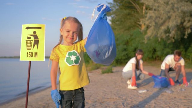 cleaning coast, portrait of little happy volunteer girl in rubber gloves with trash bag near pointer sign on background of parents collecting plastic garbage on river beach