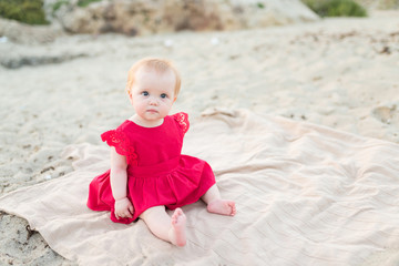 Cute baby girl in a red dress sitting and playing on the beach with sand