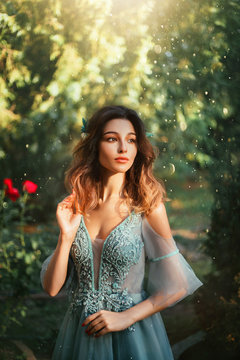tender girl with cute doll fair face and short dark hair, princess in light blue dress with open shoulders, elegant lady in garden with green trees and red roses on background, art portrait.