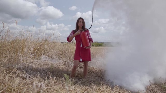 Attractive woman with long hair using fire extinguisher on the wheat field. Connection with nature, natural beauty. Harvest time. Fire hazard, fire precaution concept. Slow motion.