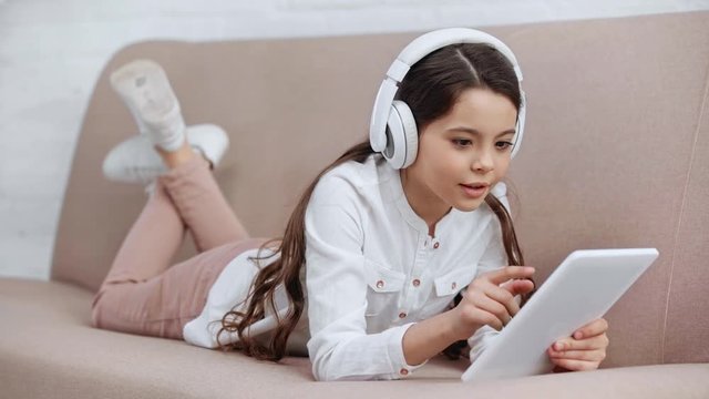cheerful preteen child lying on sofa, singing while listening music in headphones and using digital tablet