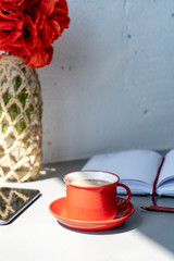 Red cup with hot coffee with milk, a vase with red poppies, a saucer, an open diary and a pencil, and a smartphone on a gray table against the background of a white wall. 