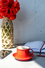 Close-up of red cup with hot coffee with milk, a vase with red poppies, a saucer, an open diary and a pencil, and a smartphone on a gray table against the background of a white wall. Vertical