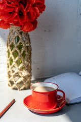 Close-up of red cup with hot coffee with milk, a vase with red poppies, a saucer, an open diary and a pencil on a gray table against the background of a white wall. Office coffee break. Vertical