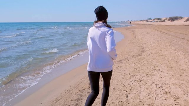 Barefooted woman is gogging on sea sand beach, back vew. Girl is wearing leggings, hoodie and hat running at sunny day. Outdoor sport activity.