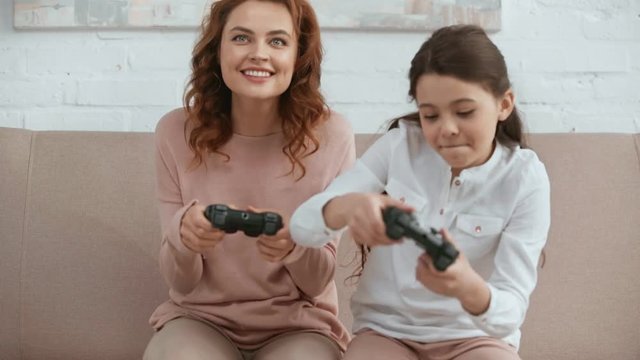 KYIV, UKRAINE -  APRIL 15, 2019: front view of mother and daughter holding joysticks and laughing while playing video game on sofa in living room