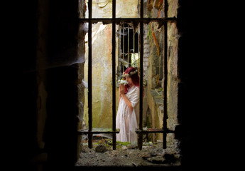 A young woman is trapped inside a jail ruin. She is  dressed in white and wears a thorn wreath on her  head. 