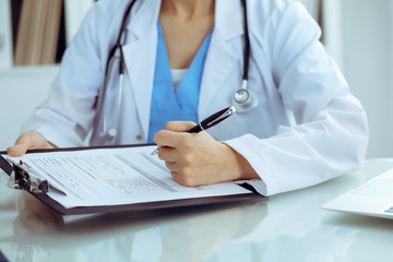 Doctor woman filling up medical form while sitting at the table, close-up of hands