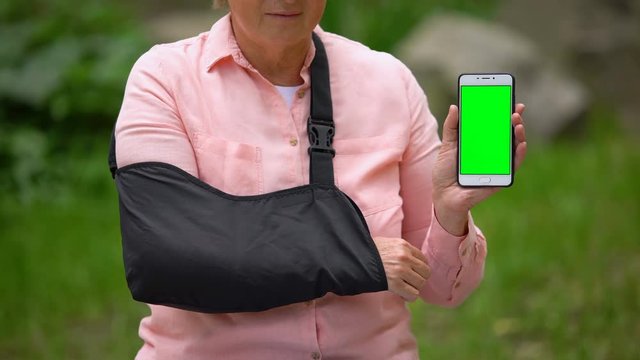 Senior woman wearing shoulder immobilizing sling showing smartphone green screen