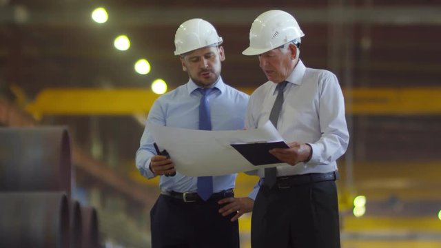 Thigh-up two shot of senior grey-haired factory executive discussing technical scheme with middle-aged production unit manager, both wearing white hardhats, against steel pipes in background