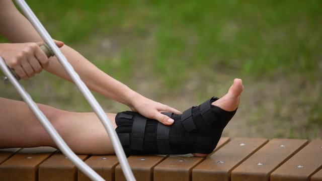 Female with crutch sitting bench with ankle brace on leg, bone fracture, strain