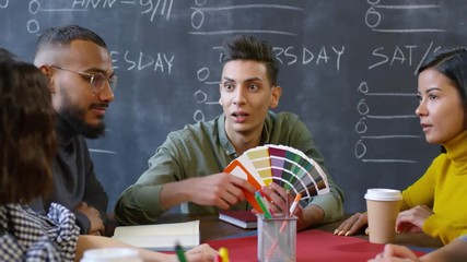 Panning chest-up shot of young creative designers collaborating on project, sitting together at table during brainstorming meeting, with one Asian guy demonstrating colour chart for others to discuss