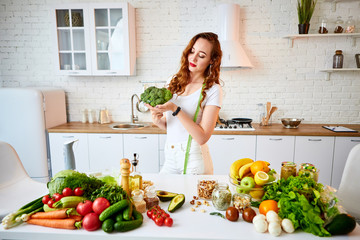 Young happy woman holding broccoli in the beautiful kitchen with green fresh ingredients indoors. Healthy food and Dieting concept. Loosing Weight