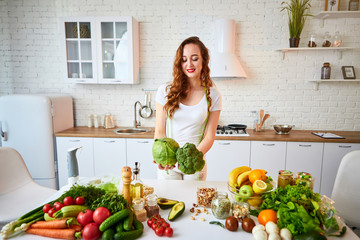 Young happy woman holding broccoli and cabbage in the beautiful kitchen with green fresh ingredients indoors. Healthy food and Dieting concept. Loosing Weight