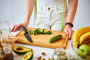 Young happy woman cutting cucumber for making salad in the beautiful kitchen with green fresh ingredients indoors. Healthy food and Dieting concept. Loosing Weight