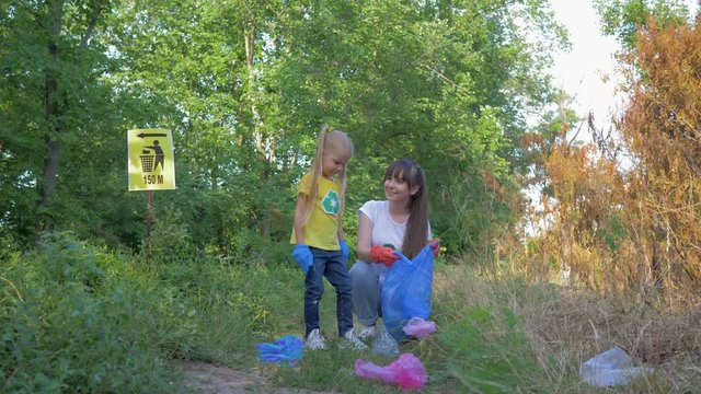 ecological care, little child girl helps mom to collect plastic and polyethylene litter in garbage bag while cleaning nature near pointer sign in green grass outdoors