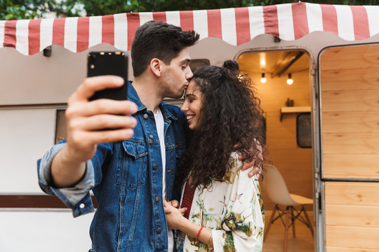 Image of man kissing pretty woman and taking selfie on cellphone near house on wheels outdoors