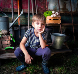 Cute emotional little boy posing against the background of an old barn wall and garden tools in low key style
