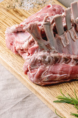 Raw uncooked rack of lamb on wooden cutting board with salt, herbs rosemary, pepper on grey linen table cloth as background.
