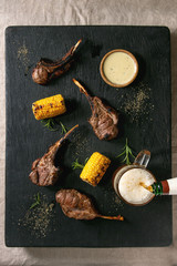 Fototapeta na wymiar Grilled bbq rack of lamb with sweet corn cobs, rosemary and cheese sauce, glass of lager beer, flowing from bottle, on black burned wooden cutting board on linen cloth background. Flat lay, space