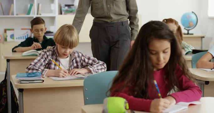 Caucasian children writing their exam in the copybooks during lesson at school and teacher walking among desks, then coming to one boy and helping him.