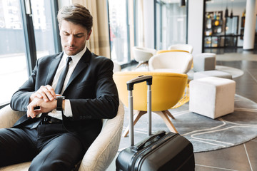Photo of successful young businessman looking at his wristwatch while sitting in hotel hall with suitcase
