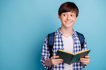 Portrait of charming boy holding printed fiction book smiling dressed in checked shirt rucksack isolated over blue background