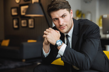 Portrait of confident young businessman wearing formal black suit sitting on armchair in hotel hall