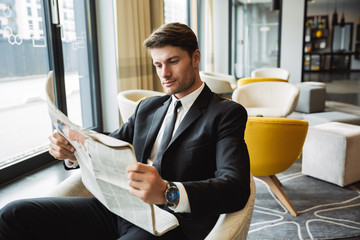 Portrait of confident young businessman sitting on armchair and reading newspaper in hotel hall